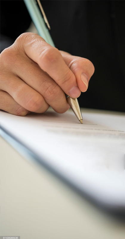 a male wearing a black suite uses his right hand signs a piece of white paper with a pen